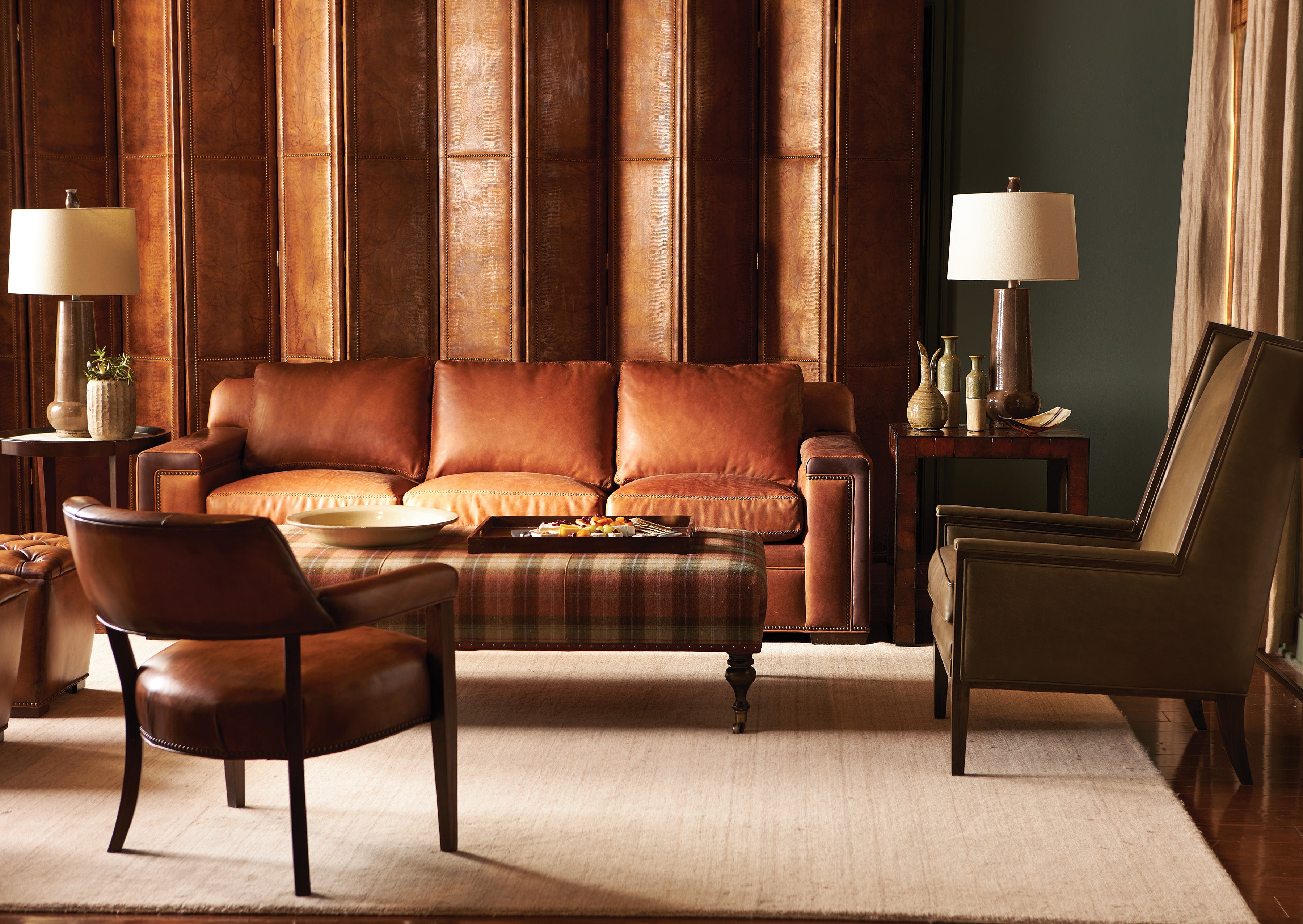 A living room showcasing what appears to be a Hancock & Moore Artemis Sofa in brown leather, paired with a brown leather side chair, an upholstered armchair with piping details, a plaid upholstered ottoman with dark wooden legs, and two side tables with lamps against a backdrop of textured brown wall panels and a dark green wall.