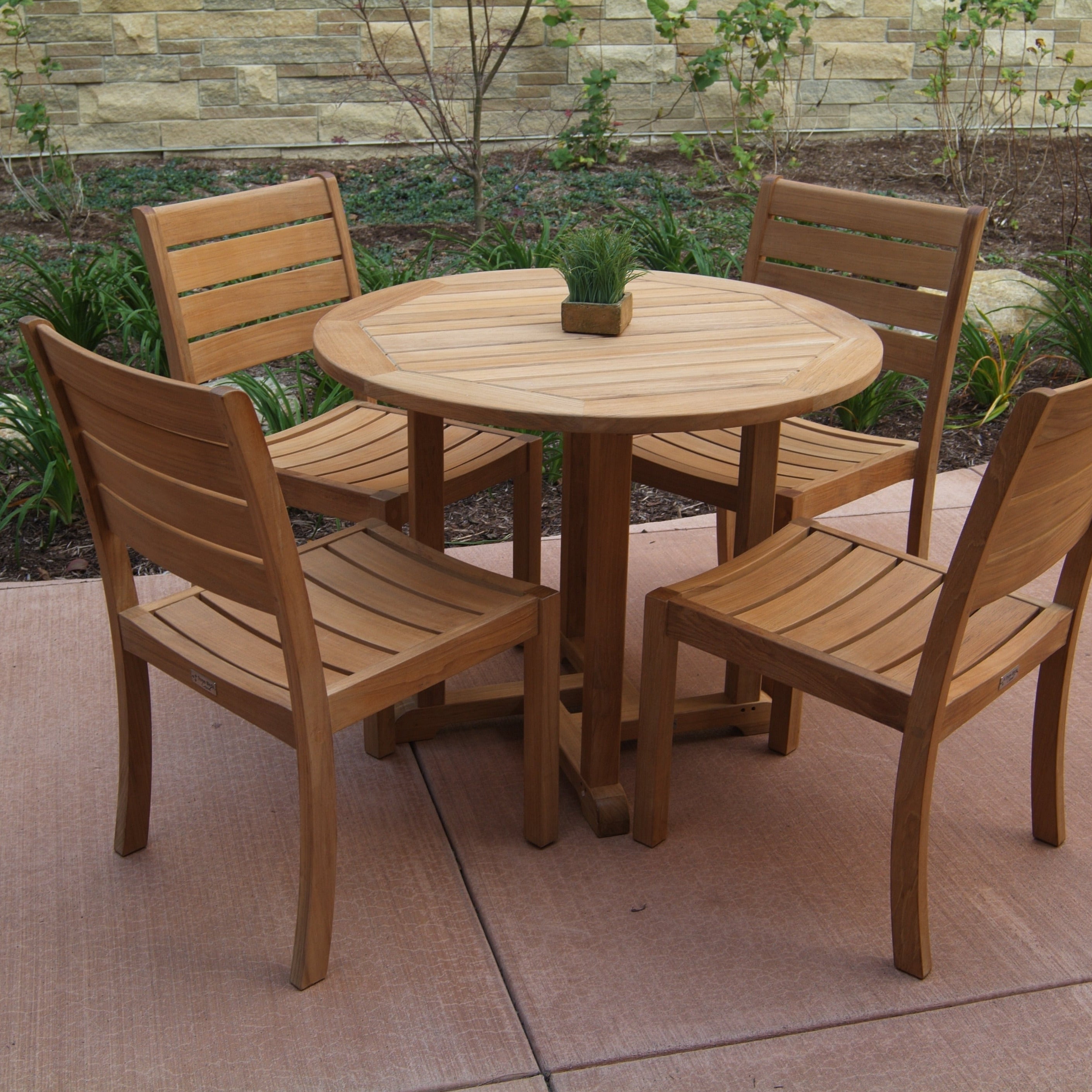 A Three Birds Oxford Teak Round 42" Dining Table set with four matching chairs on a stone patio, with greenery and a stone wall in the background.