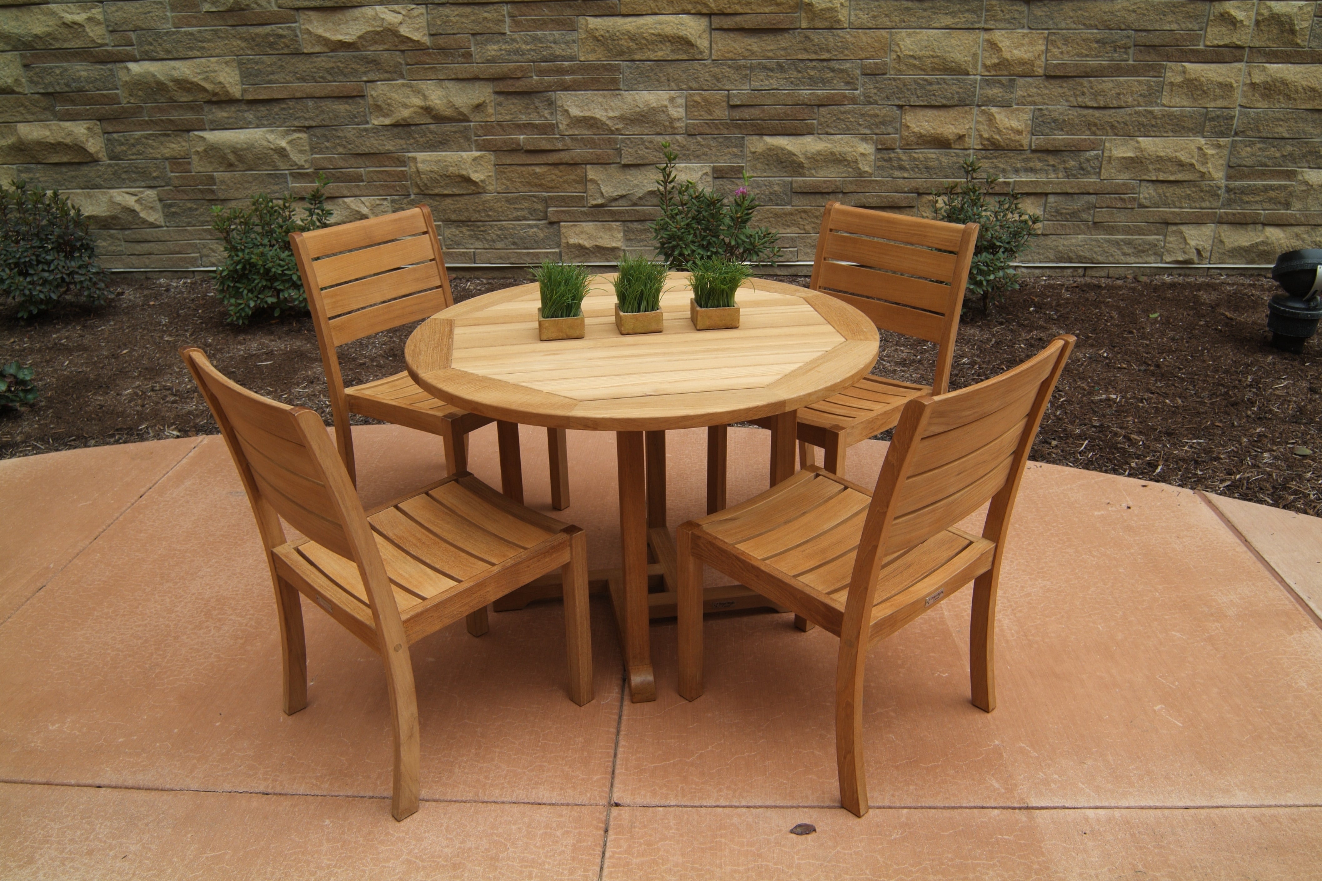 A Three Birds Oxford Teak Round 42" Dining Table set with four matching chairs on a stone patio, with greenery and a stone wall in the background.