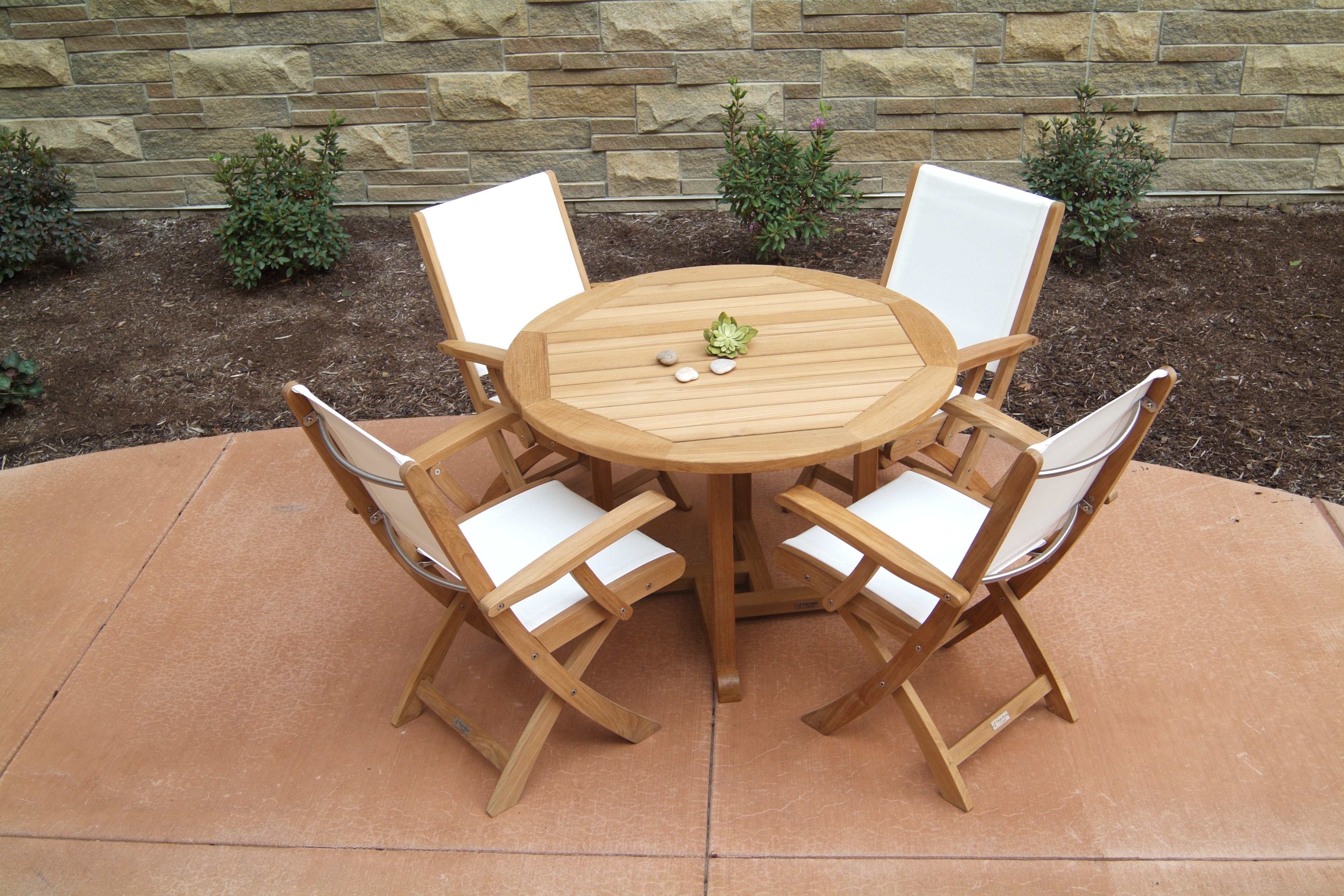 A Three Birds Oxford Teak Round 42" Dining Table set with four matching chairs on a stone patio, with greenery and a stone wall in the background.