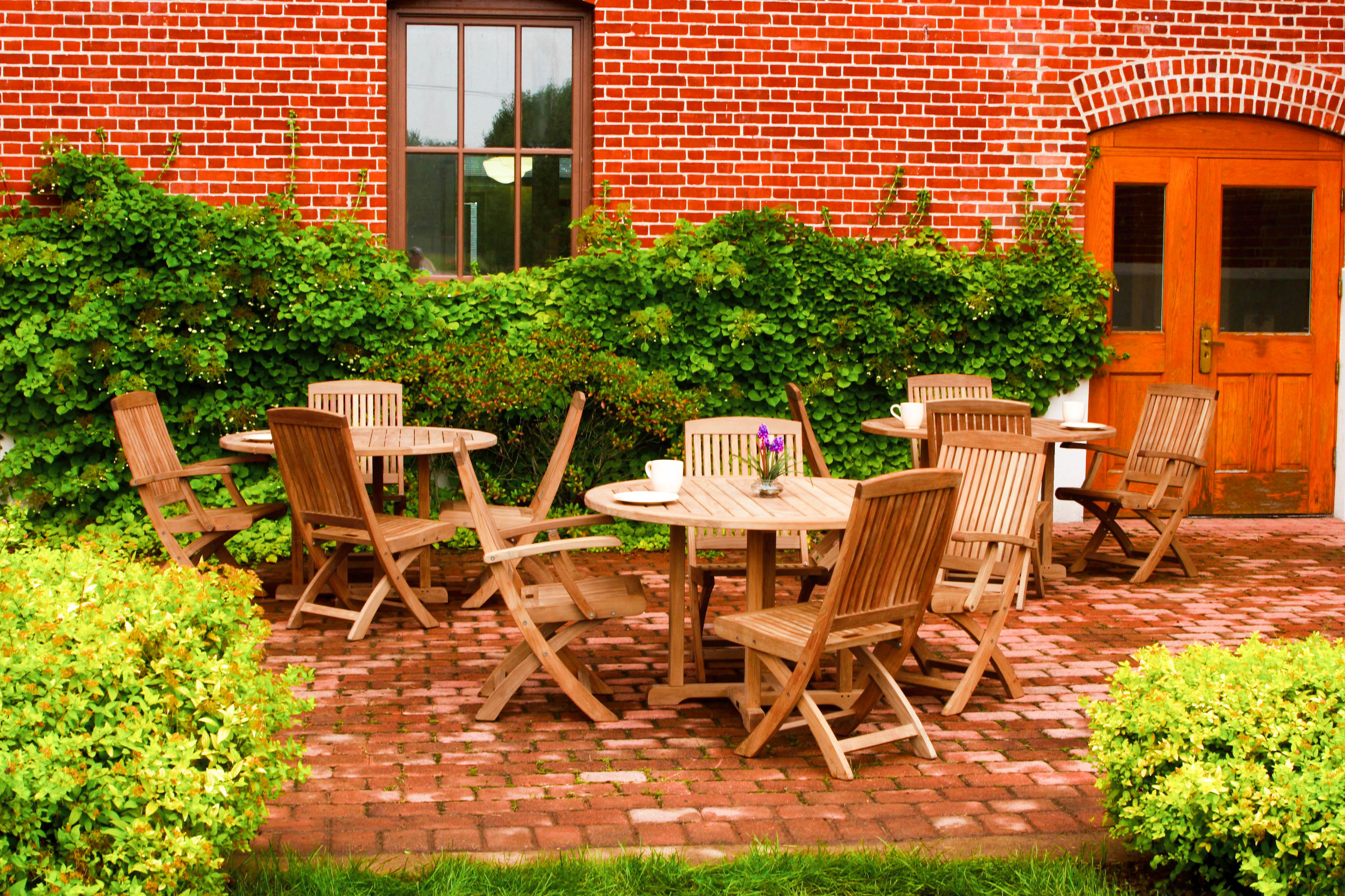 A Three Birds Oxford Teak Round 42" Dining Table set with four matching chairs on a stone patio, with greenery and a stone wall in the background.