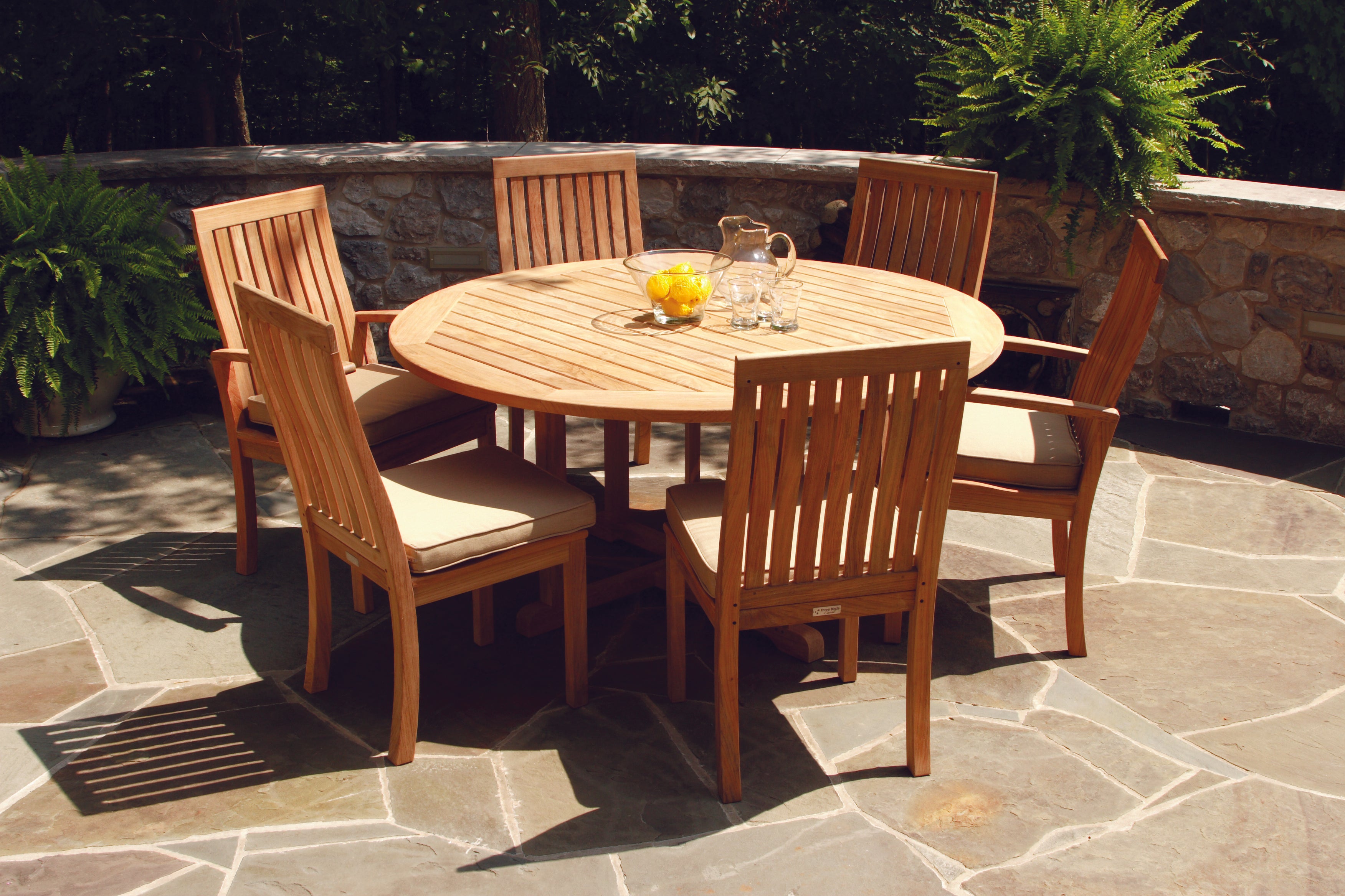 A Three Birds Oxford 60" 7-Piece Dining Set shown in Knoxville, Tennessee, featuring a large round teak table with a slatted top and a pedestal base, surrounded by six Oxford dining chairs with vertical slats and light beige cushions, set on a flagstone patio with a low stone wall, ferns, and trees in the background.