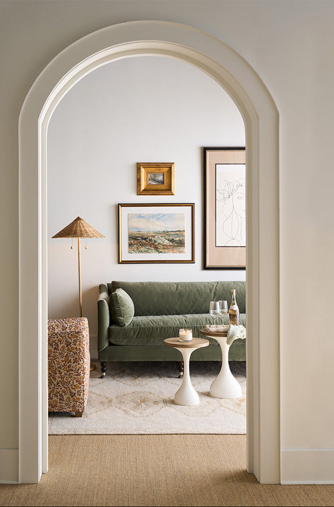 Living room with a green sofa, framed artwork, and a lamp, viewed through an archway.