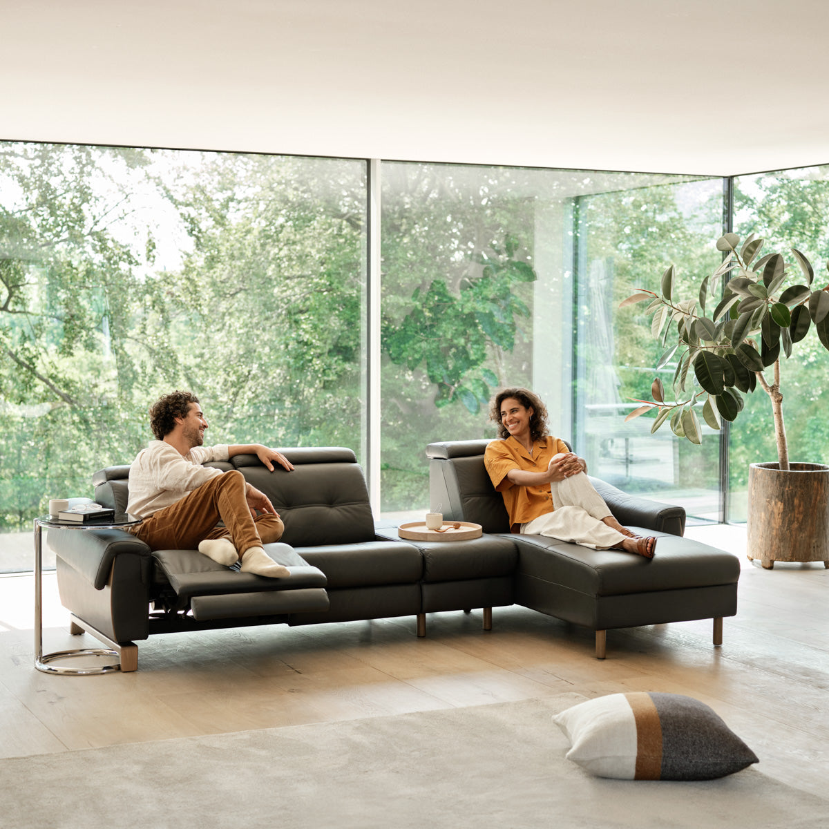 Two people sitting on a modern sectional sofa in a bright living room with large windows.